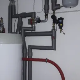 Instalacja grzewcza w kotłowni: biały zbiornik, szare rury z izolacją, czerwona i niebieska rura giętka, manometr na zbiorniku wyrównawczym, zawory i filtry, widoczne ślady po wierceniu w ścianie.