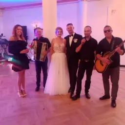Zespół muzyczny z Jaktorowa pozuje do zdjęcia z parą młodą w eleganckiej sali weselnej, akordeon, gitara akustyczna i mikrofony w tle.
