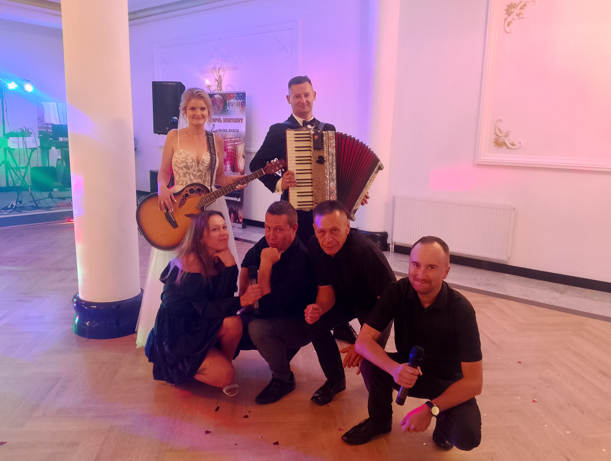 Zespół muzyczny pozuje do zdjęcia: kobieta z gitarą, mężczyzna z akordeonem i trzech wokalistów z mikrofonami, w tle dekoracje sali weselnej.