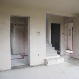 Wnętrze w trakcie remontu: surowe ściany po tynkowaniu maszynowym z widocznymi instalacjami elektrycznymi i fragmentem schodów w stanie deweloperskim.