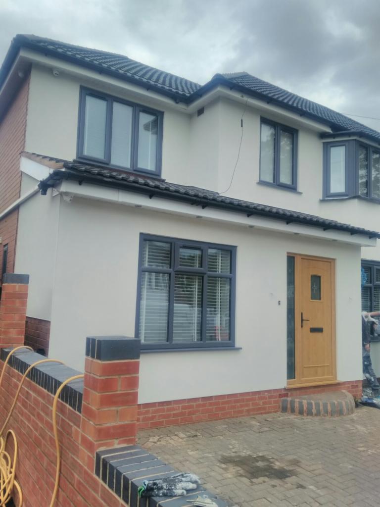 Dom jednorodzinny po remoncie elewacji, z nowymi oknami w grafitowych ramach i świeżo otynkowaną, jasną elewacją. Widoczny fragment murku z cegły klinkierowej w kolorze czerwonym z grafitowymi...