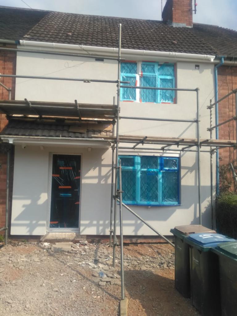 Dom w trakcie prac elewacyjnych; okna i drzwi zabezpieczone folią, widoczne rusztowania, świeżo nałożony tynk w kolorze jasnoszarym.