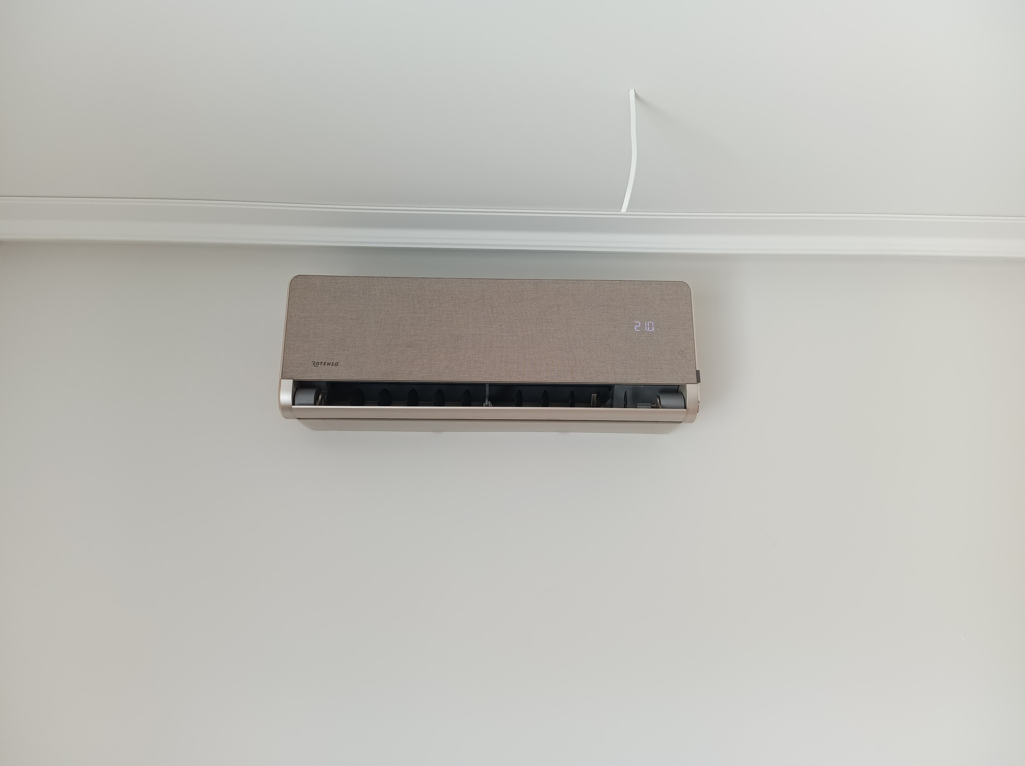 Beżowa klimatyzacja Rotenso w minimalistycznym wnętrzu, zamontowana na białej ścianie pod sufitem z listwą dekoracyjną. Widoczny przewód zasilający.