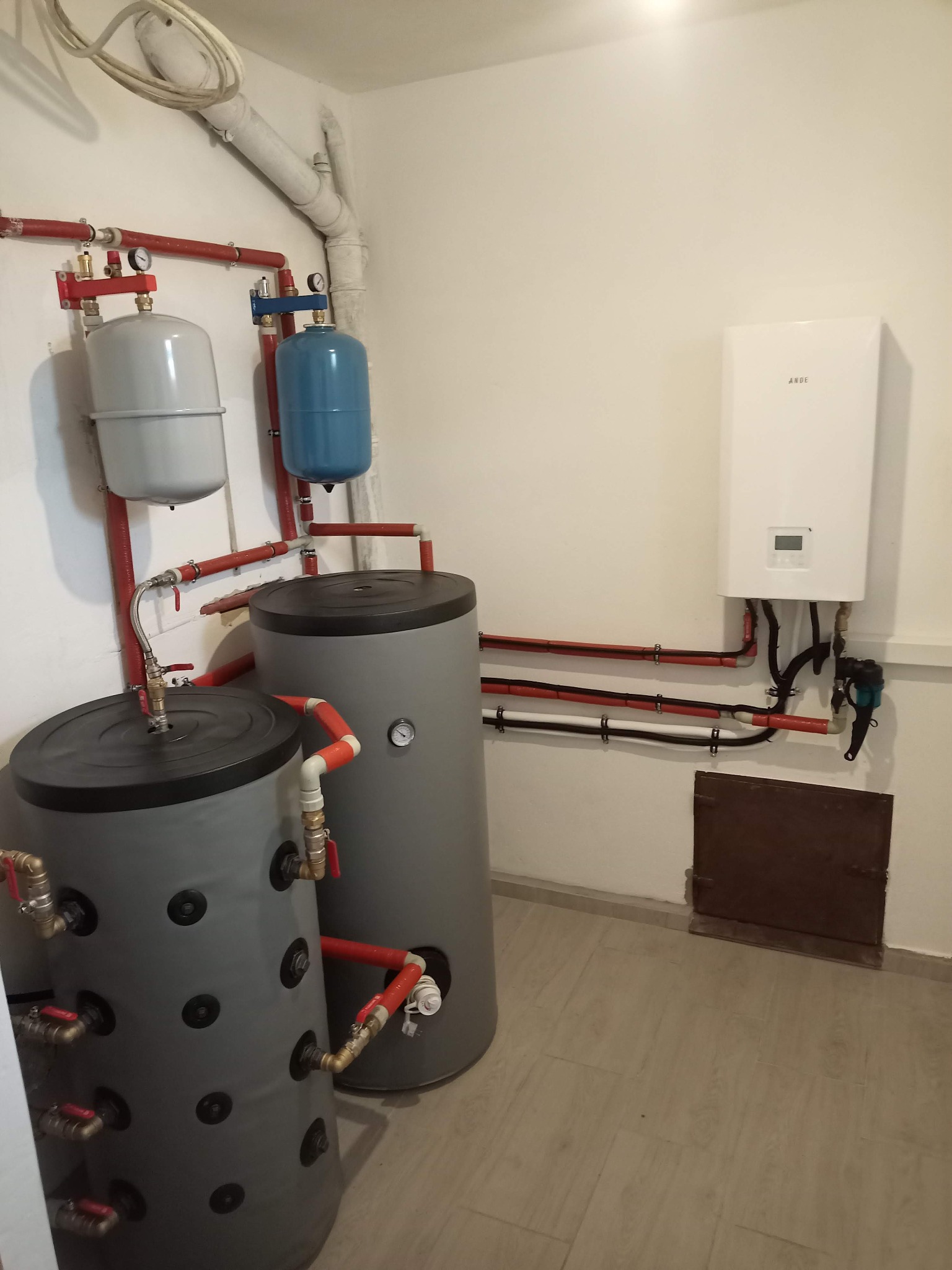 Instalacja grzewcza w kotłowni: dwa szare zbiorniki akumulacyjne, biały piec gazowy na ścianie, czerwone rury i białe rury. Elementy instalacji połączone ze sobą.