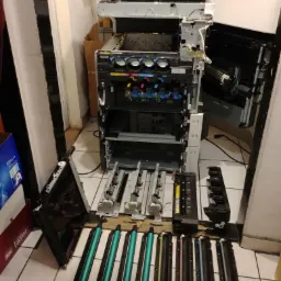 Rozłożona na części kserokopiarka z widocznymi elementami wewnętrznymi i wymienionymi tonerami, ustawiona na podłodze z płytek.