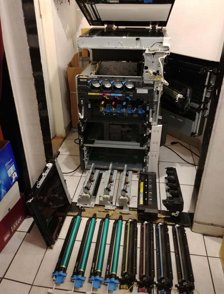 Rozłożona na części kserokopiarka z widocznymi elementami wewnętrznymi i wymienionymi tonerami, ustawiona na podłodze z płytek.