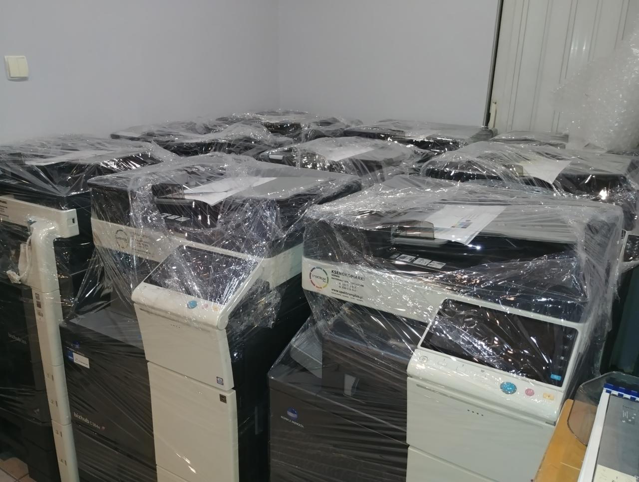 Nowe kserokopiarki zabezpieczone folią, gotowe do transportu w Krakowie. Widoczne modele Bizhub C554e i podobne.