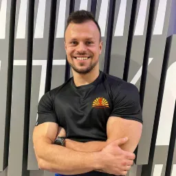 Uśmiechnięty mężczyzna z założonymi rękami, ubrany w czarną koszulkę z logo 'Forma na lata', stojący na tle ściany z geometrycznym wzorem w odcieniach szarości i bieli.