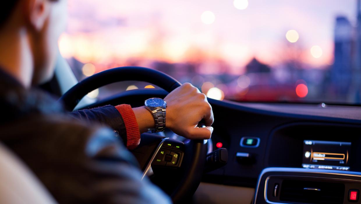 Kierowca z zegarkiem na ręku trzyma kierownicę w samochodzie, w tle rozmyte światła miasta o zmierzchu.