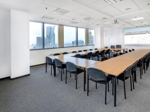 Jasna sala konferencyjna z układem stołów w kształcie litery U, widok na miasto przez okna, rzutnik na suficie i tablica interaktywna.