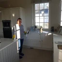 Kobieta w żółtych rękawicach sprząta białą kuchnię z widokiem na dom przez okno, używając sprayu i ściereczki.