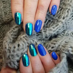 Dłonie z paznokciami pomalowanymi na dwa kolory: intensywny niebieski oraz opalizujący zielono-niebieski, na tle szarego, grubego swetra.