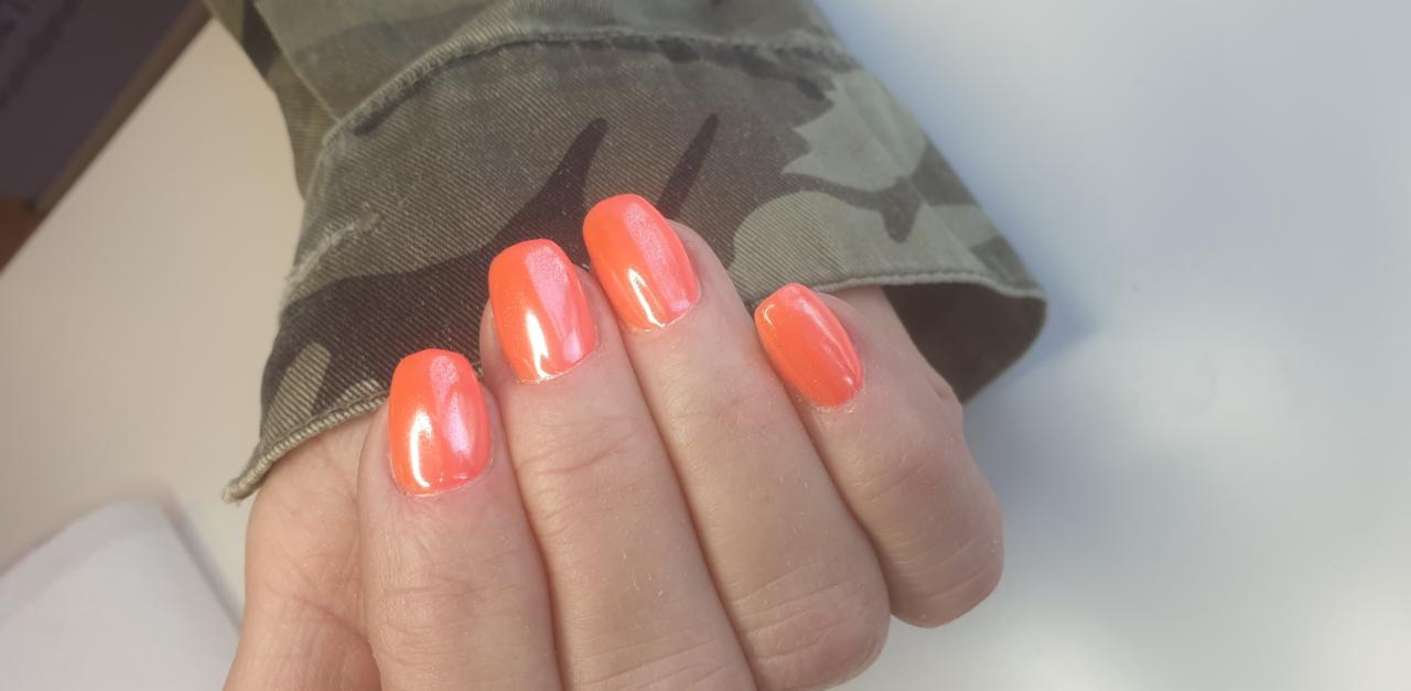 Pomarańczowe paznokcie o owalnym kształcie z delikatnym połyskiem, na dłoni kobiety w kurtce moro.