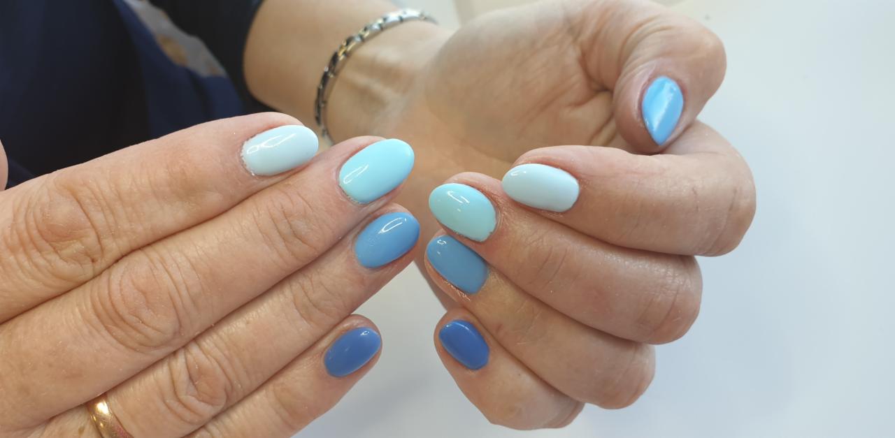 Dłonie kobiety z owalnymi paznokciami pomalowanymi lakierem w odcieniach błękitu, od jasnego pastelowego po ciemny, królewski błękit, na białym tle, widoczny pierścionek na palcu.