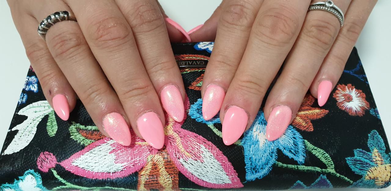 Ręce z pomalowanymi na różowo paznokciami w kształcie migdała, błyszczące wykończenie, na tle torebki z haftowanymi kwiatami.