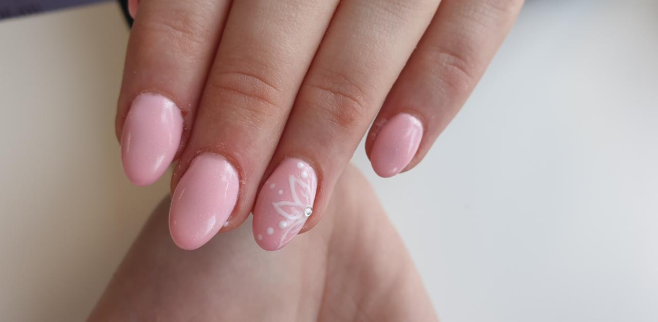 Dłoń z pomalowanymi na jasny różowo owalnymi paznokciami. Na jednym z paznokci biały kwiat z cyrkonią pośrodku i białymi kropkami.
