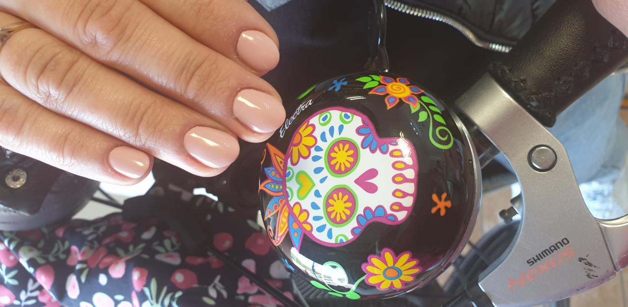 Świeżo pomalowane, beżowe paznokcie na dłoni kobiety, obok ozdobnego dzwonka rowerowego z motywem czaszki i kwiatów.