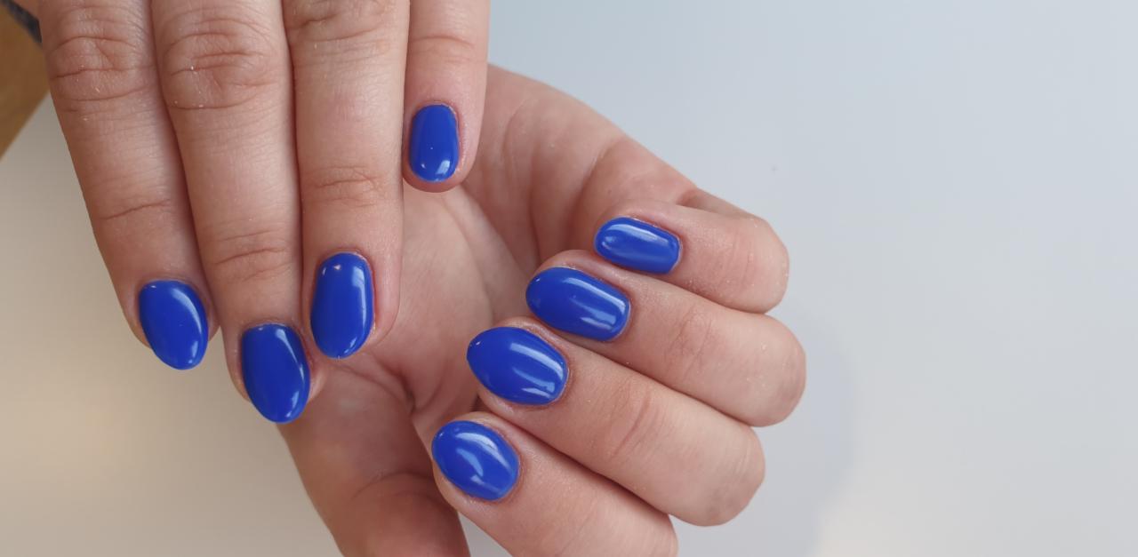 Dłonie z owalnymi paznokciami pokrytymi jednolitym, intensywnym, kobaltowym lakierem hybrydowym na jasnym tle.