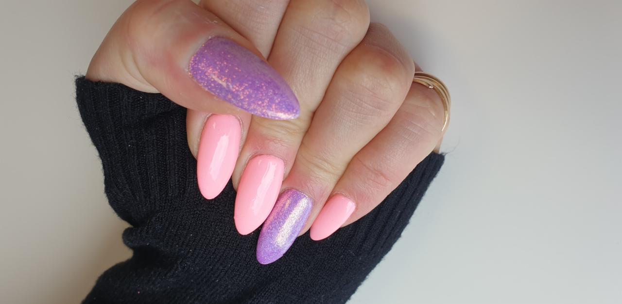 Dłoń z pomalowanymi paznokciami w odcieniach różu i fioletu z brokatowym wykończeniem, na tle czarnego swetra i białego blatu.