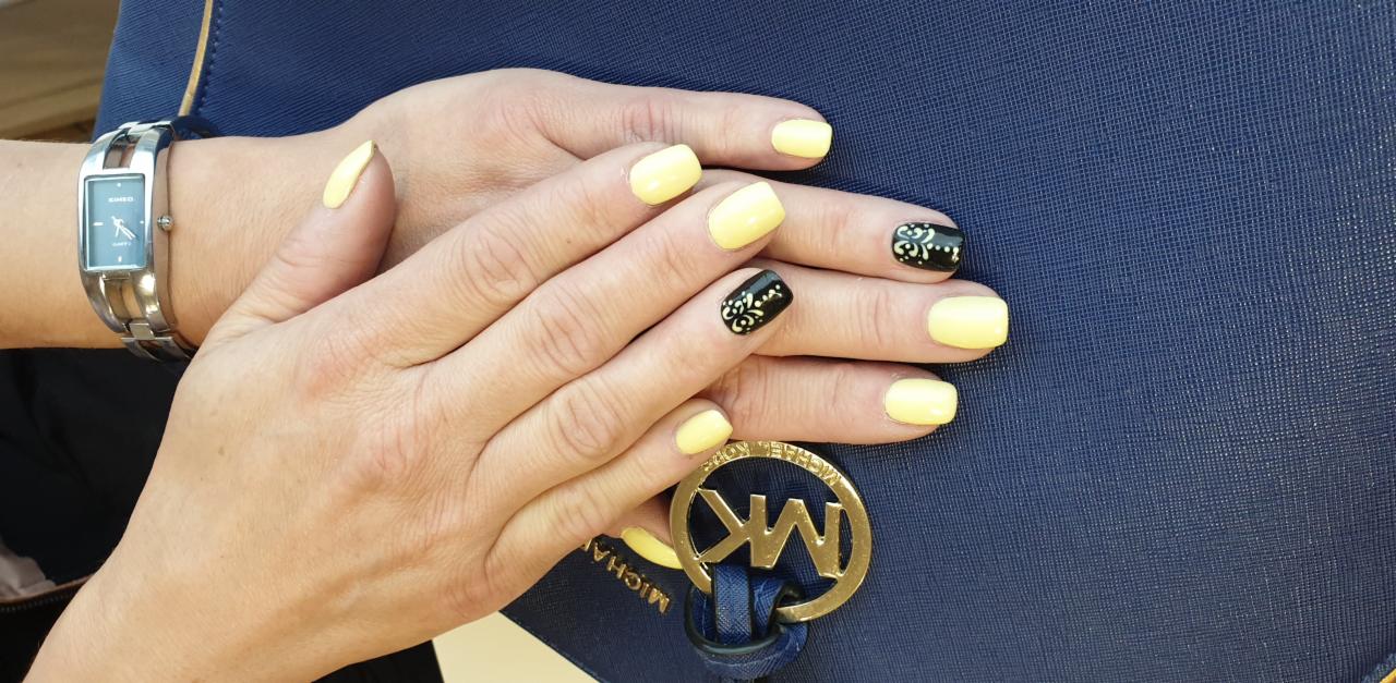 Dłonie z żółtym manicurem i czarnymi akcentami zdobniczymi na tle granatowej torebki z logo Michael Kors i srebrnym zegarkiem Casio.