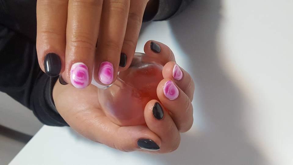 Dłonie z pomalowanymi paznokciami, na niektórych paznokciach wzór marmurkowy w odcieniach różu i bieli, na innych ciemny lakier, trzymające okrągły pojemnik z płynem.