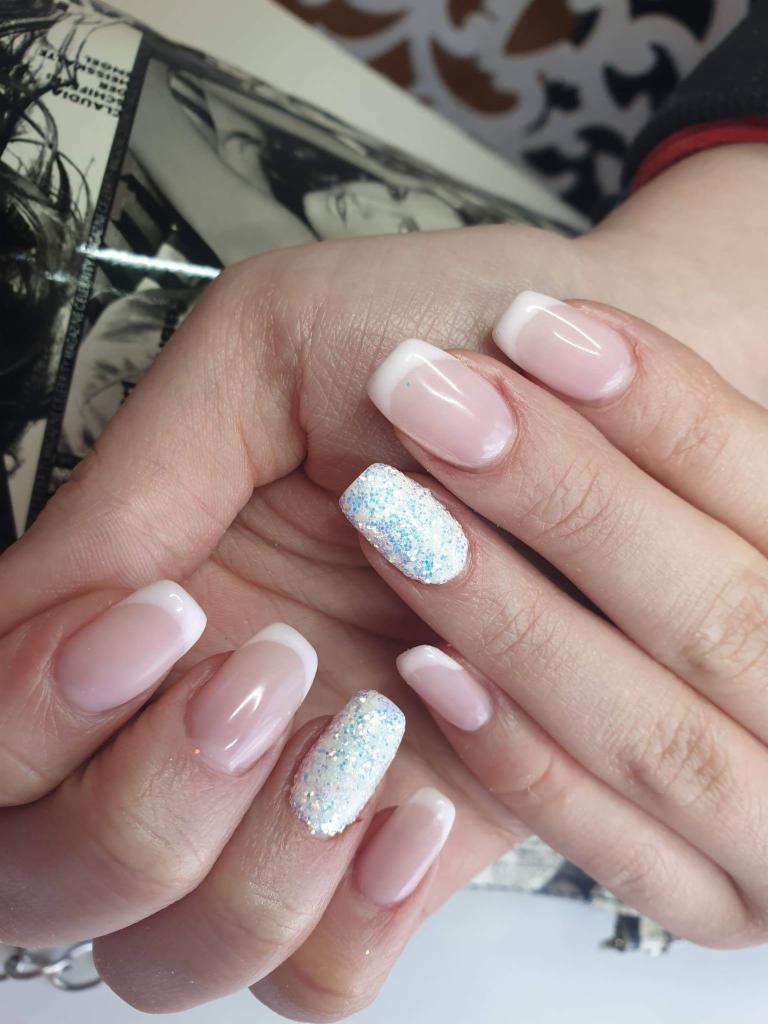 Dłonie z manicure hybrydowym: klasyczny french na większości paznokci, dwa paznokcie pokryte białym brokatem z niebieskimi refleksami, zdjęcie wykonane w świetle dziennym.