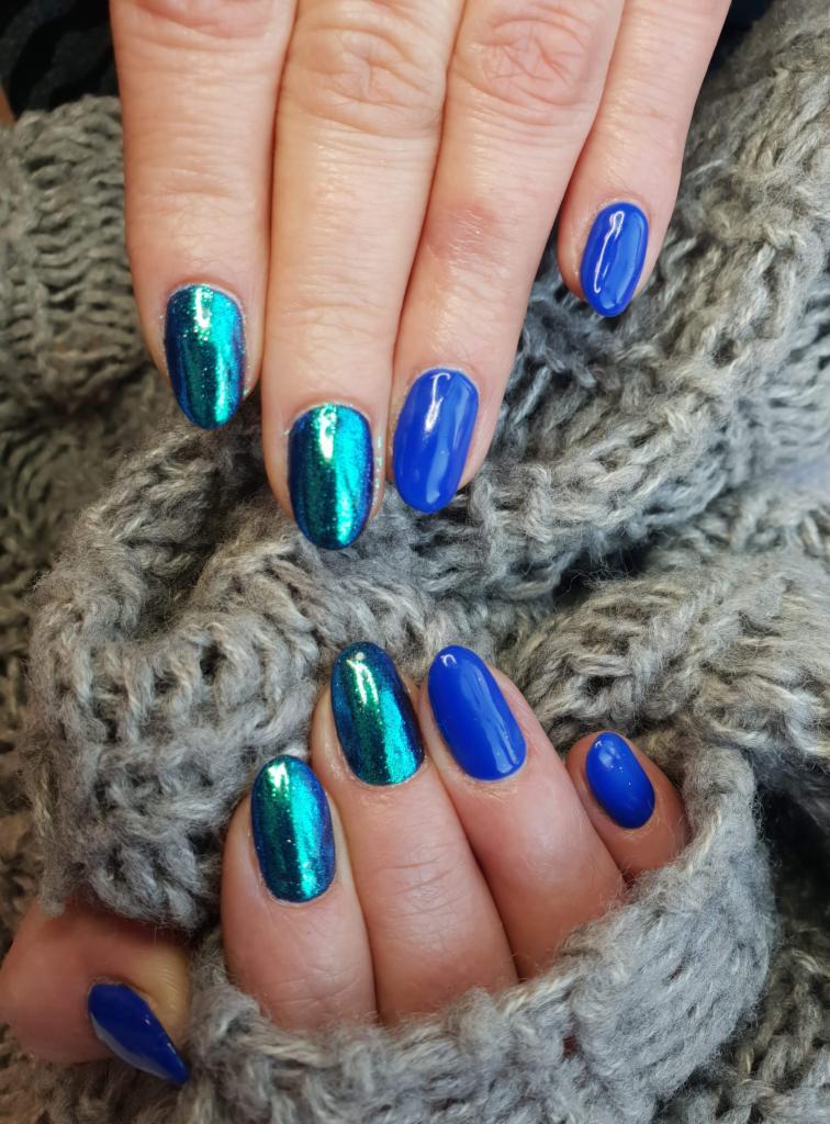Dłonie z paznokciami pomalowanymi na dwa kolory: intensywny niebieski oraz opalizujący zielono-niebieski, na tle szarego, grubego swetra.