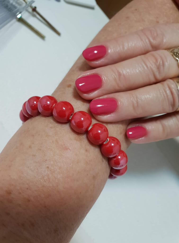 Prawa dłoń z świeżo pomalowanymi paznokciami w odcieniu malinowej czerwieni, na nadgarstku czerwona bransoletka z okrągłych korali, w tle narzędzia do manicure.