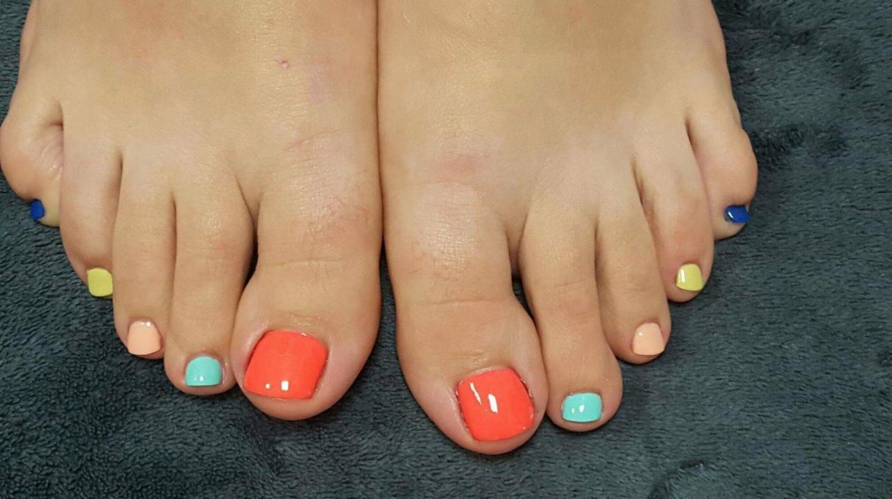 Zbliżenie na stopy z pomalowanymi paznokciami u stóp w różnych pastelowych kolorach: koralowym, turkusowym, brzoskwiniowym, żółtym i niebieskim, na szarym ręczniku.
