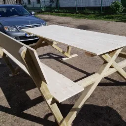 Surowy drewniany stół piknikowy z ławkami, konstrukcja krzyżakowa, jasne drewno, na ziemi, z zaparkowanym srebrnym Mercedesem w tle.