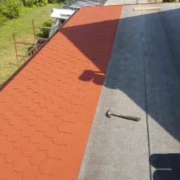 Częściowo pokryty dach papą i gontem bitumicznym w kolorze ceglastym, młotek leżący na papie, rusztowanie i zieleń w tle.