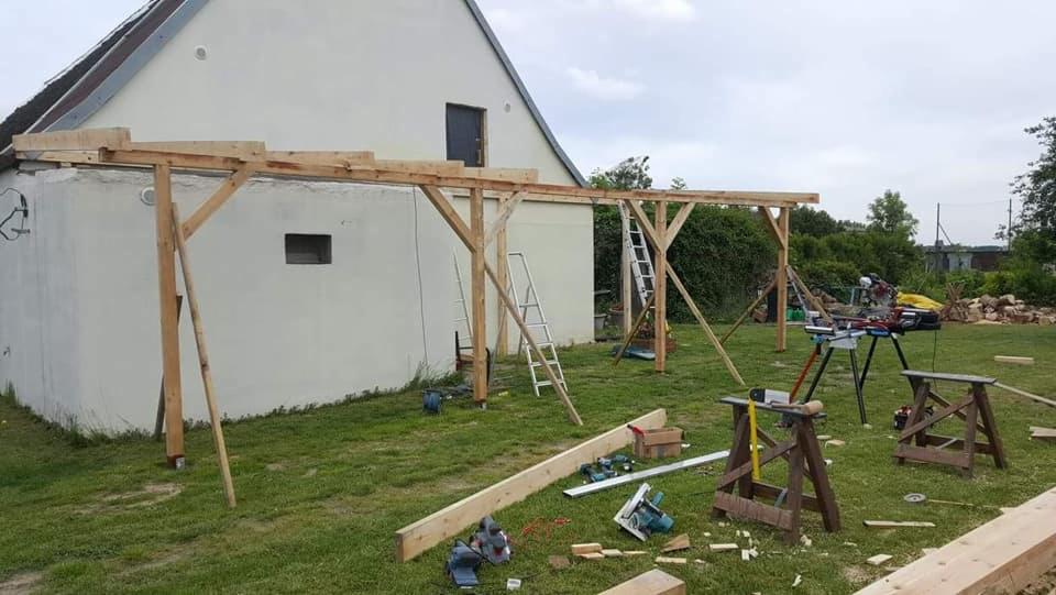 Konstrukcja drewniana zadaszenia w trakcie budowy, widoczne słupy, belki, drabiny, piły elektryczne i stemple, na trawniku przed budynkiem.