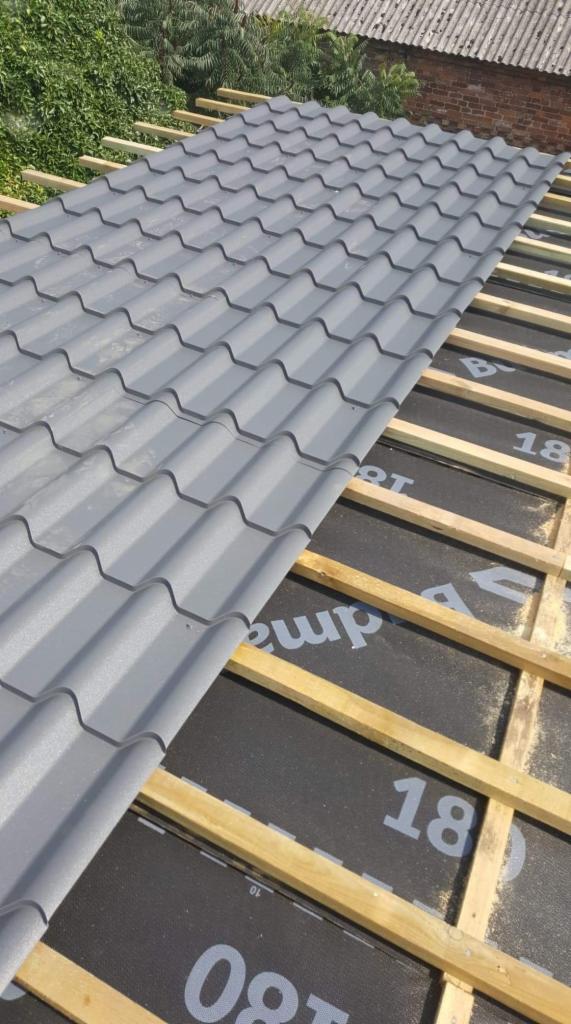 Fragment dachu z szarej blachodachówki ułożonej na drewnianej konstrukcji i membranie dachowej z widocznym nadrukiem, w tle zieleń i mur z cegły.