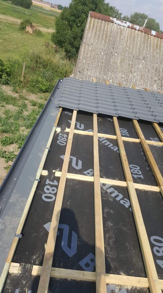 Szary dach z blachy trapezowej w trakcie montażu, widoczna membrana dachowa z nadrukiem i drewniana konstrukcja łatowania, w tle budynek z eternitu i zielona łąka.