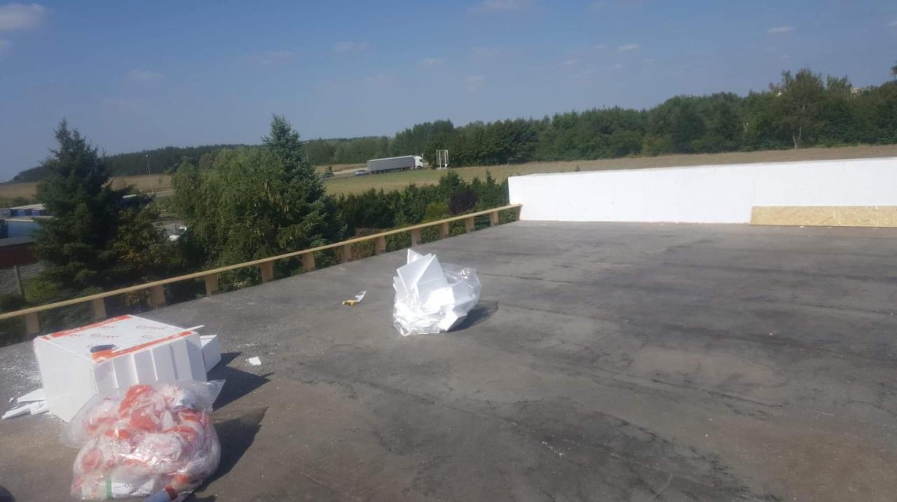 Płaski dach pokryty papą, z widocznymi materiałami izolacyjnymi (styropian) w worku i pudełkach, drewniana barierka na krawędzi, w tle drzewa i przejeżdżająca ciężarówka.