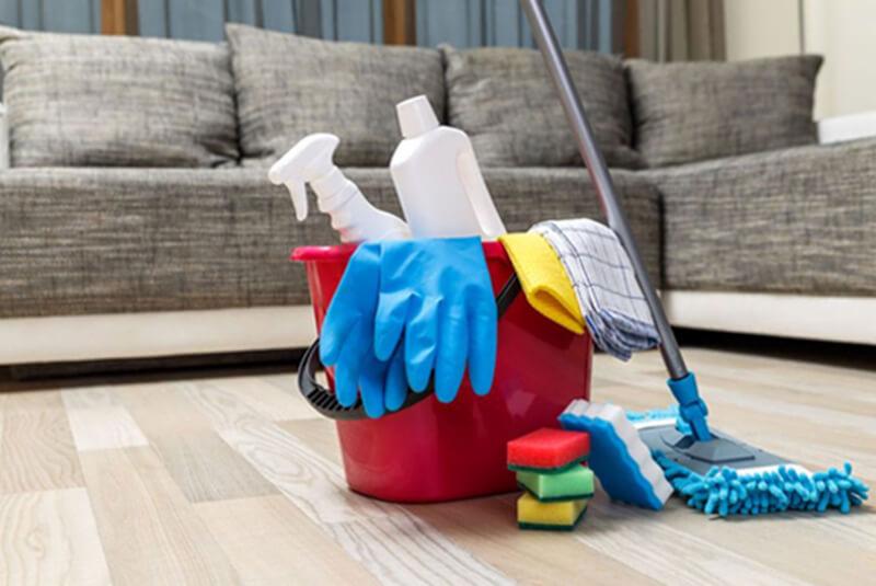 Czerwone wiadro z niebieskimi rękawicami, ściereczkami i środkami czystości stoi obok mopa na drewnianej podłodze, w tle szara kanapa.