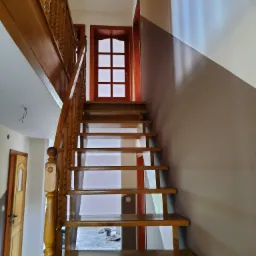 Drewniane schody z kręconą balustradą, prowadzące na górę, ściana pomalowana na dwa kolory, zabezpieczona podłoga folią malarską.