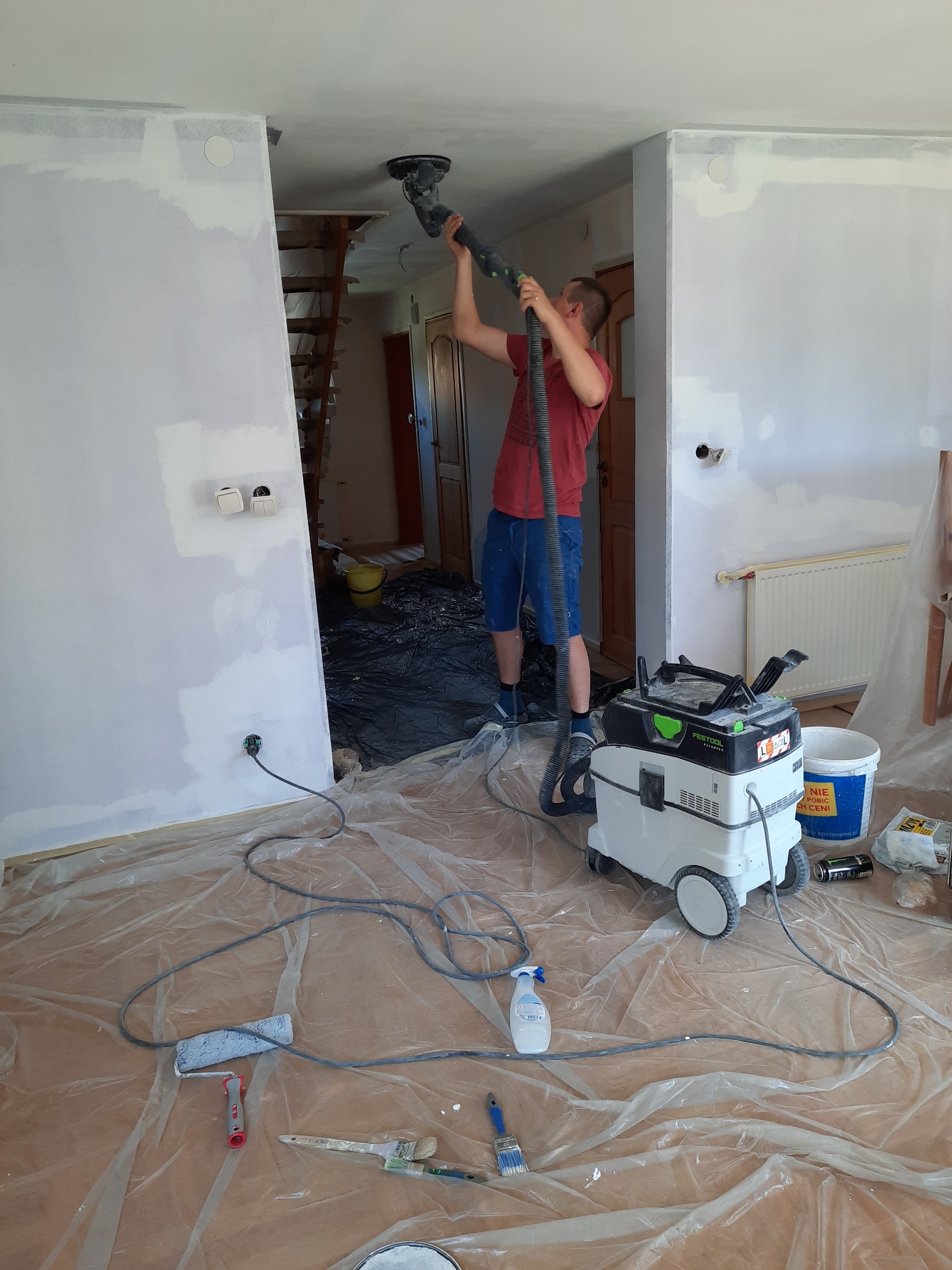 Mężczyzna szlifuje sufit za pomocą szlifierki z odkurzaczem Festool, przygotowując powierzchnię do dalszych prac wykończeniowych; podłoga zabezpieczona folią, widoczne narzędzia malarskie.