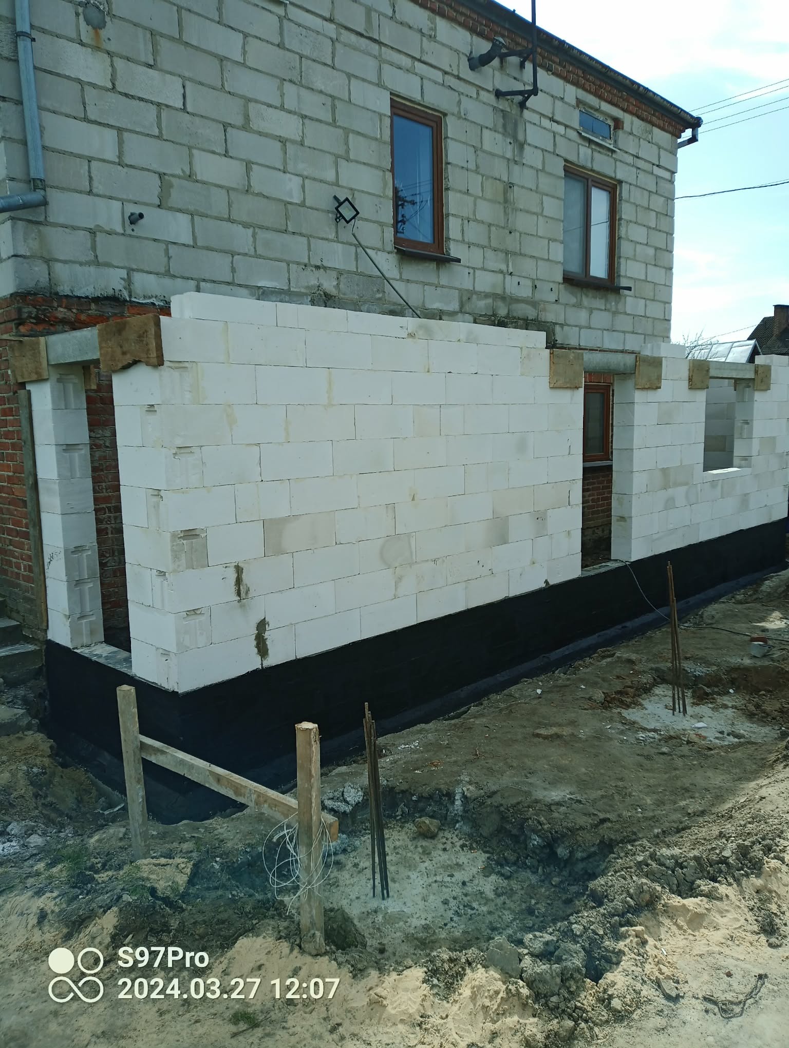 Wznoszenie ściany z białych bloczków na fundamencie z izolacją przeciwwilgociową. Widoczne okna w istniejącej części budynku. Prace murarskie w toku.