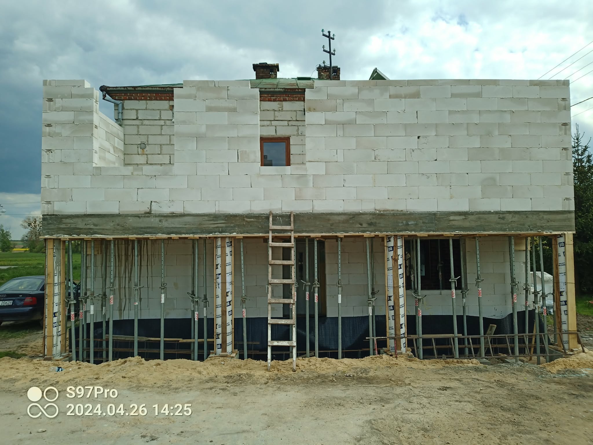 Wznoszenie ścian domu z białych bloczków, widoczne podpory szalunkowe i drabina. Częściowo zrealizowana konstrukcja z oknem i otworami na instalacje.