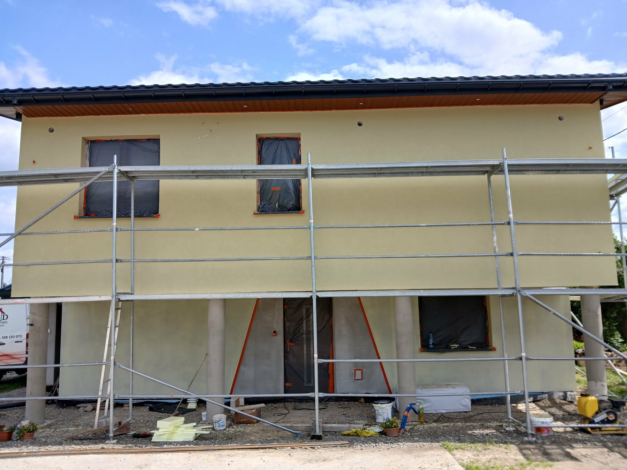 Elewacja domu w trakcie prac wykończeniowych, z nałożonym jasnym tynkiem i rusztowaniem. Okna i drzwi zabezpieczone folią. Widoczne materiały budowlane i narzędzia.