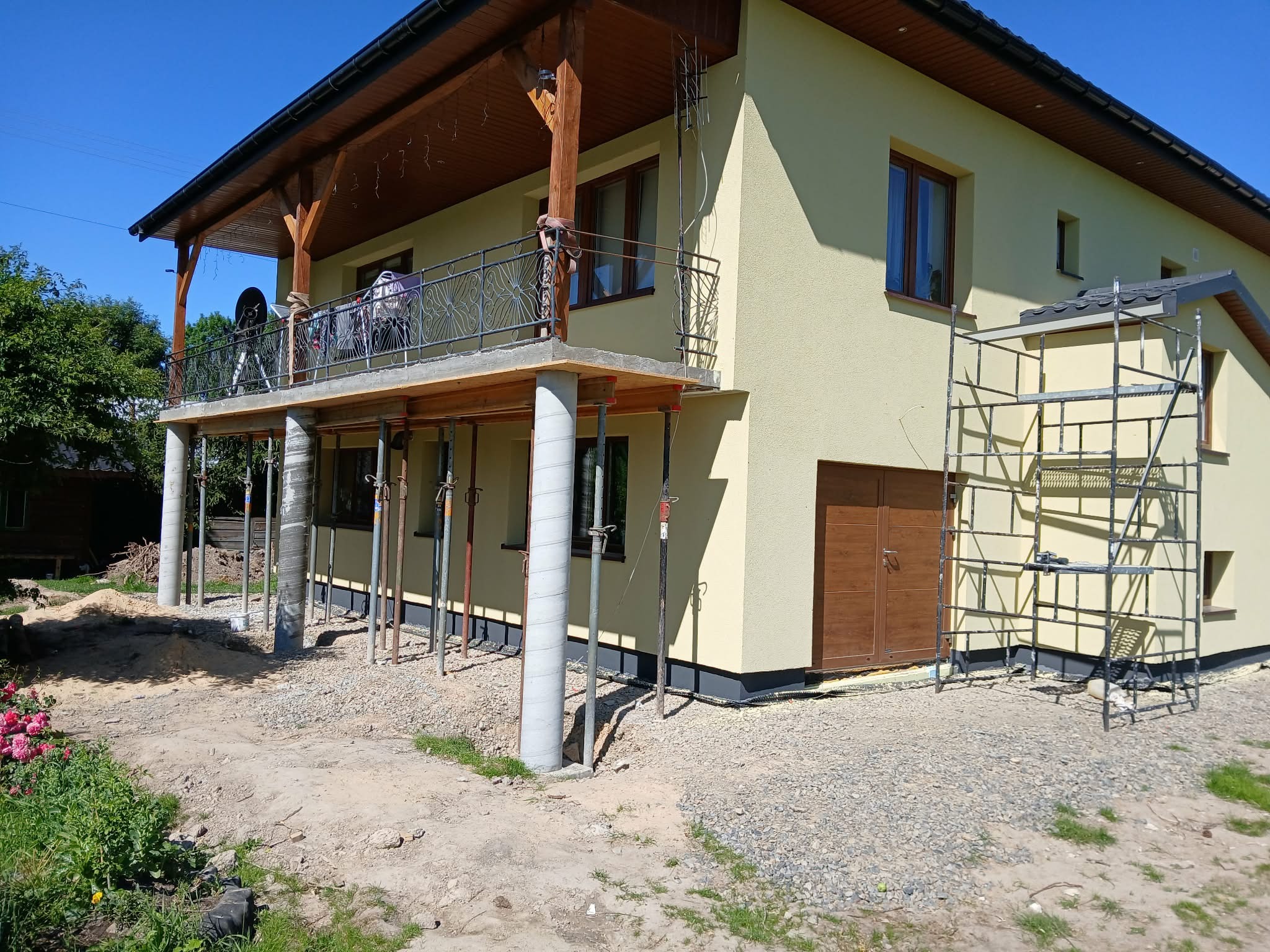 Żółty dom z balkonem wspartym na betonowych kolumnach, widoczne rusztowanie i teren w trakcie prac wykończeniowych. Ujęcie w słoneczny dzień.