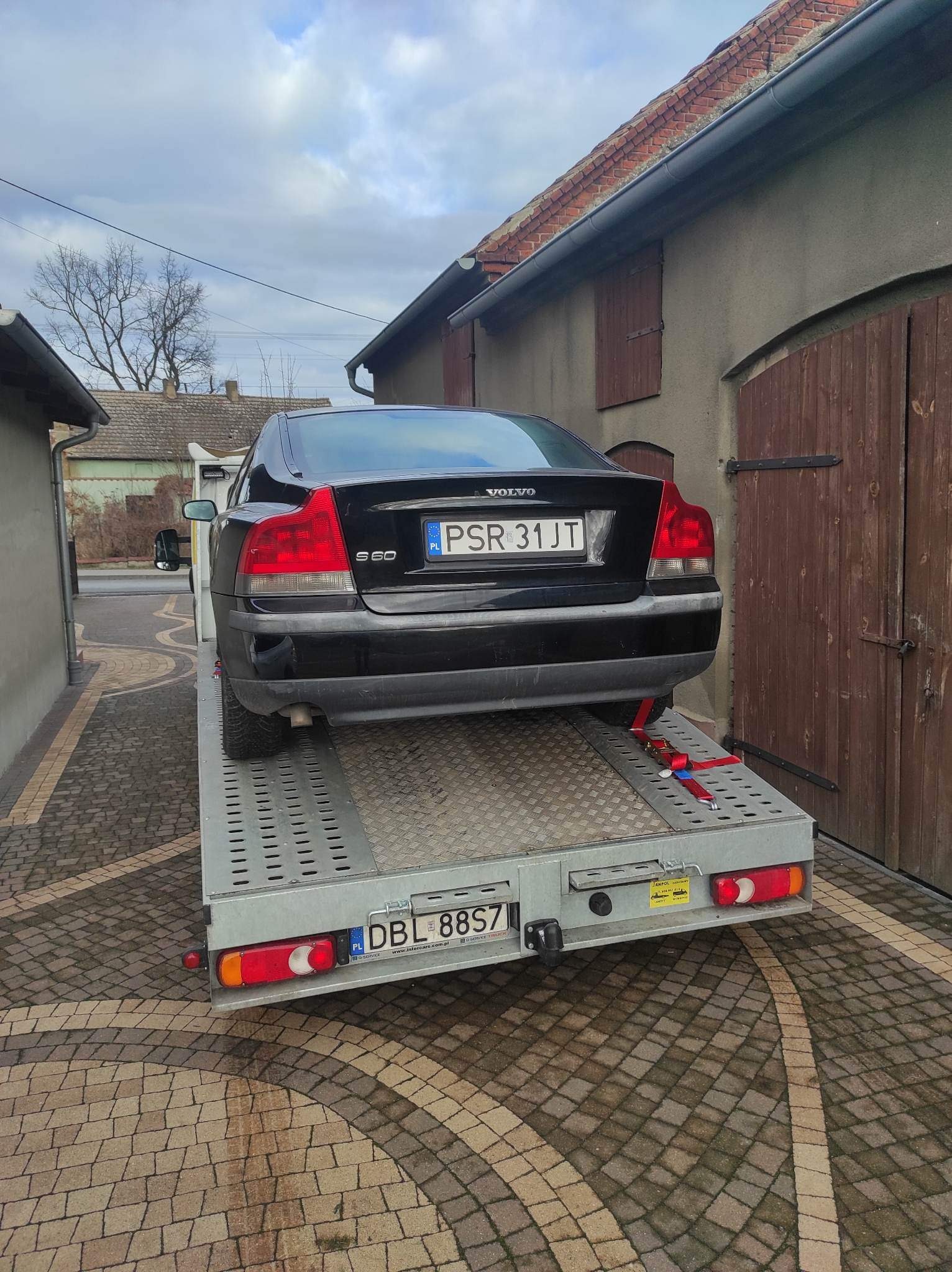 Czarny samochód Volvo S60 z polską rejestracją PSR 31JT zabezpieczony czerwonymi pasami na szarej platformie lawety, zaparkowanej na podjeździe wyłożonym kostką.