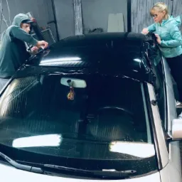 Pracownicy w warsztacie nakładają folię na dach samochodu, widoczna przednia szyba i maska auta.