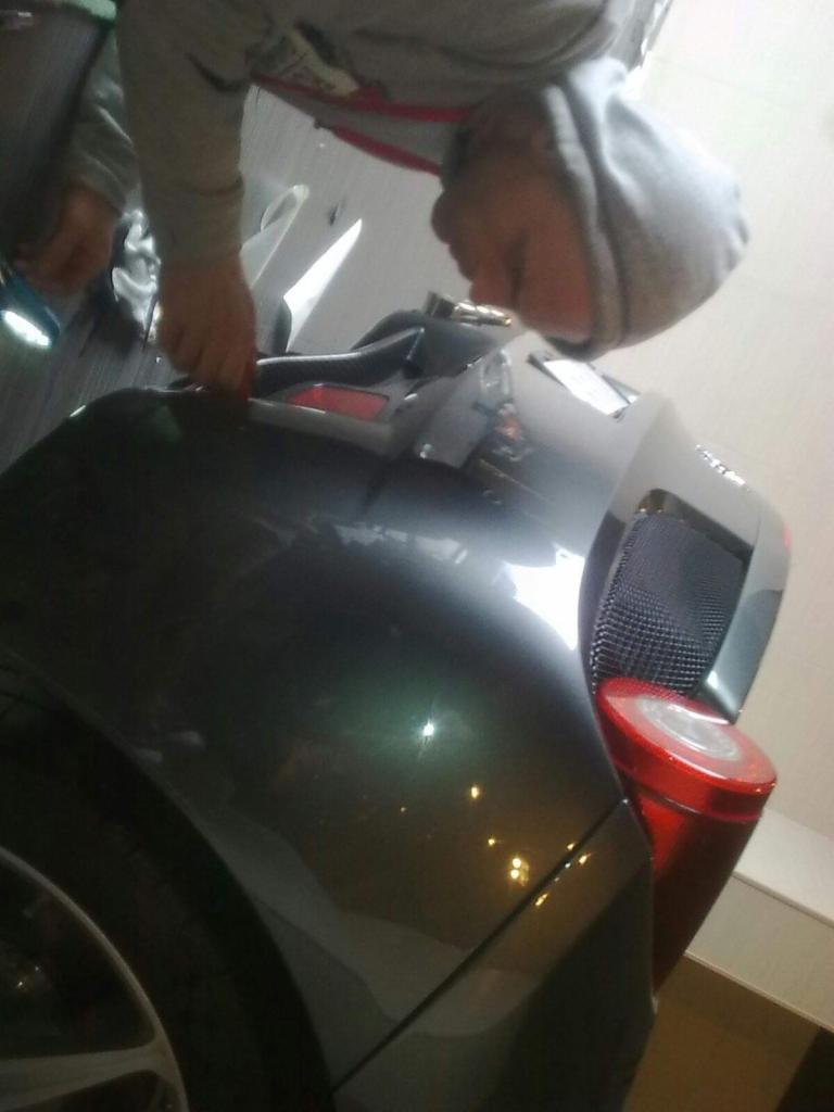 Mężczyzna w szarej bluzie z kapturem pochyla się nad tylną lampą samochodu, prawdopodobnie podczas montażu lub naprawy. Widoczne odbicia światła na karoserii.