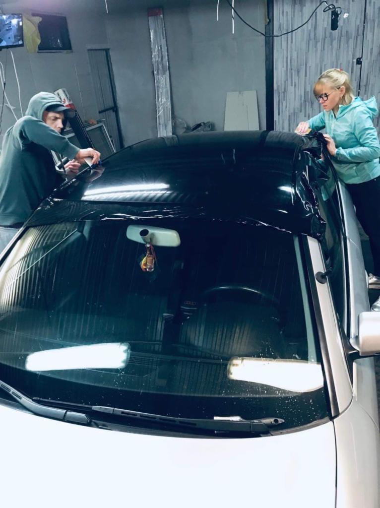 Pracownicy w warsztacie nakładają folię na dach samochodu, widoczna przednia szyba i maska auta.