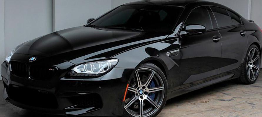 Czarny, luksusowy samochód BMW M6 Gran Coupe z przyciemnionymi szybami, zaparkowany w studiu. Widoczne sportowe felgi z niebieskimi zaciskami hamulcowymi i pomarańczowym akcentem na oponie.