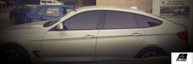 Srebrny samochód z przyciemnionymi szybami bocznymi i tylną, stojący na parkingu. W tle widoczne inne pojazdy i plakat reklamowy.