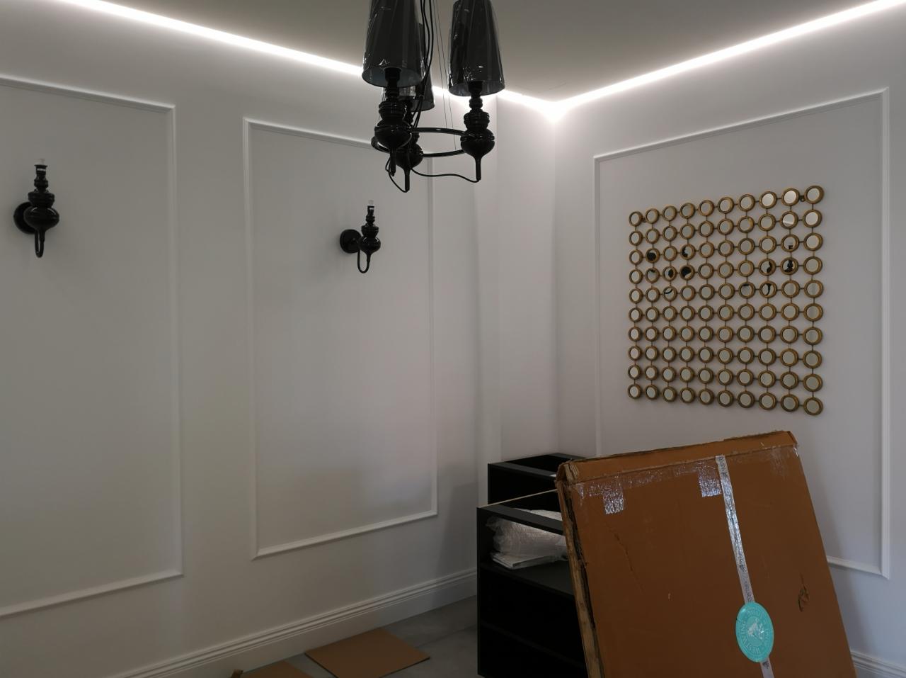 Białe ściany z dekoracyjnymi listwami i kinkietami, czarny żyrandol, ozdobna ściana z okrągłych luster, w rogu kartonowe opakowanie i czarna komoda, oświetlenie LED pod sufitem.