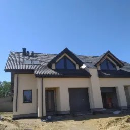 Nowoczesny dom jednorodzinny z ciemną dachówką ceramiczną i oknami dachowymi, świeżo po kryciu, widoczne prace wykończeniowe na zewnątrz budynku.